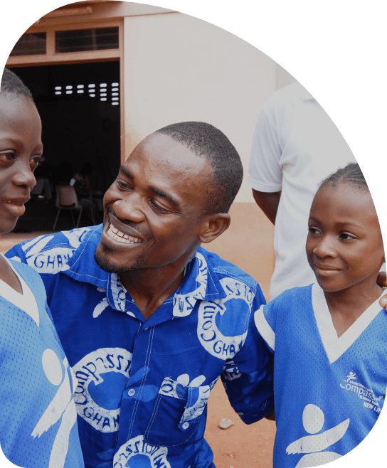 A smiling man kneels between two young children. He \ appears to be speaking to the child on his right. All \ three are wearing vibrant blue shirts.