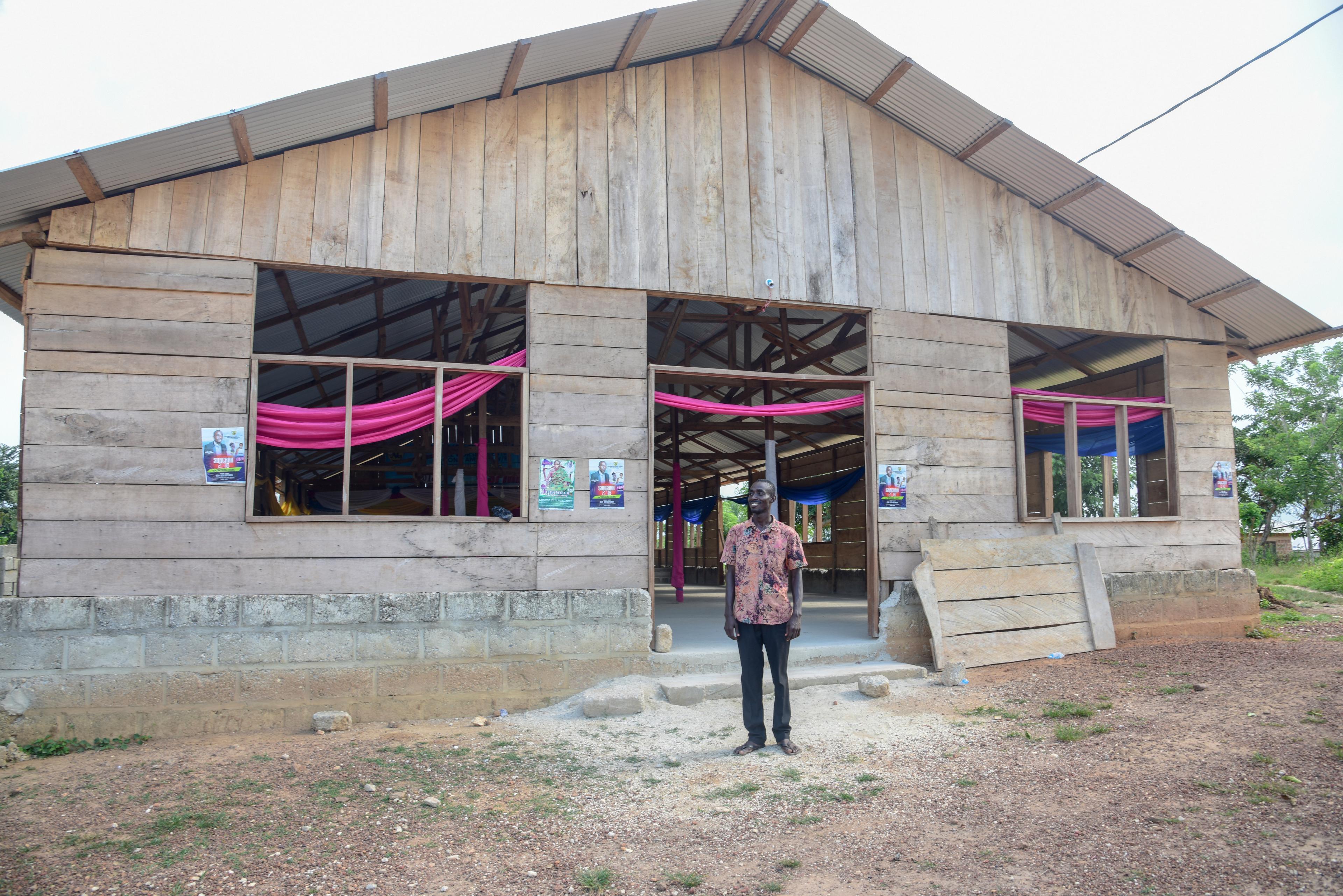 A man in black pants and a vibrant button-up shirt stands in \ front of a church building. The building has open doors and \ windows, and bright pink and blue tapestries can be seen \ hanging inside.