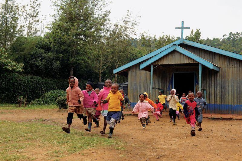 A dozen or so kids emerge running from a metal church \ building. Many of the children are bundles up in \ long-sleeved shirts or jumpers. The church is topped with \ a light blue cross.