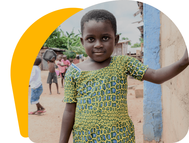 A young Ghanaian girl, maybe 5 or 6 years of age, leans \ with her left arm outstretched on a pale yellow wall. She \ makes eye contact with the camera and is wearing a yellow \ dress with traditional blue spots or patterns.