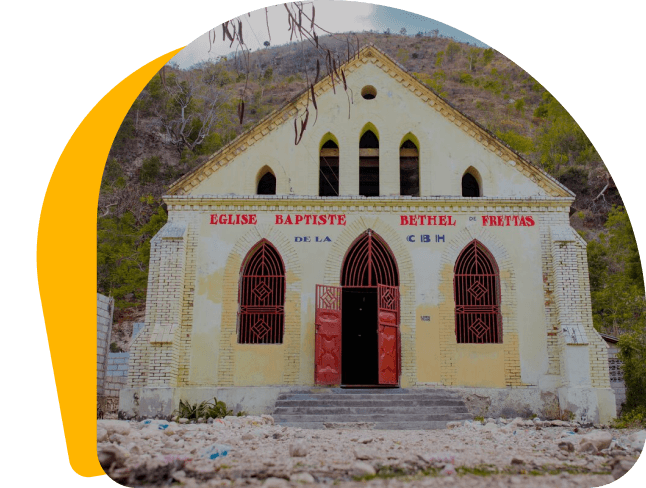 The front façade of a brick and plaster church. Its walls \ are pale yellow, contrasting with its bold red door, which \ is open, and the bright red framing around its windows.