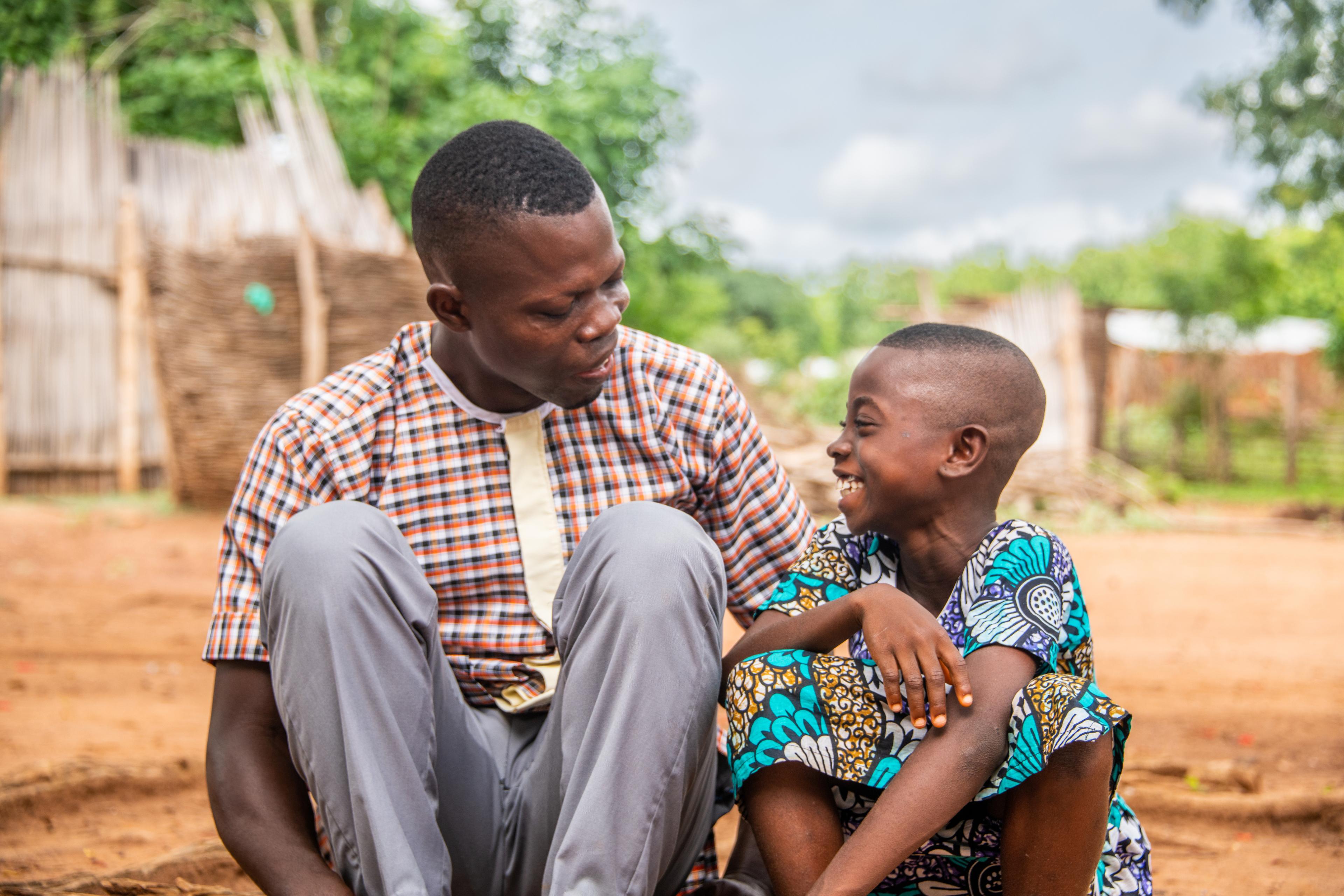 An African man seated next to a younger boy. The boy is smiling wide and is wearing a colorful, traditional outfit. A village is blurred in the background with trees and a bright sky.